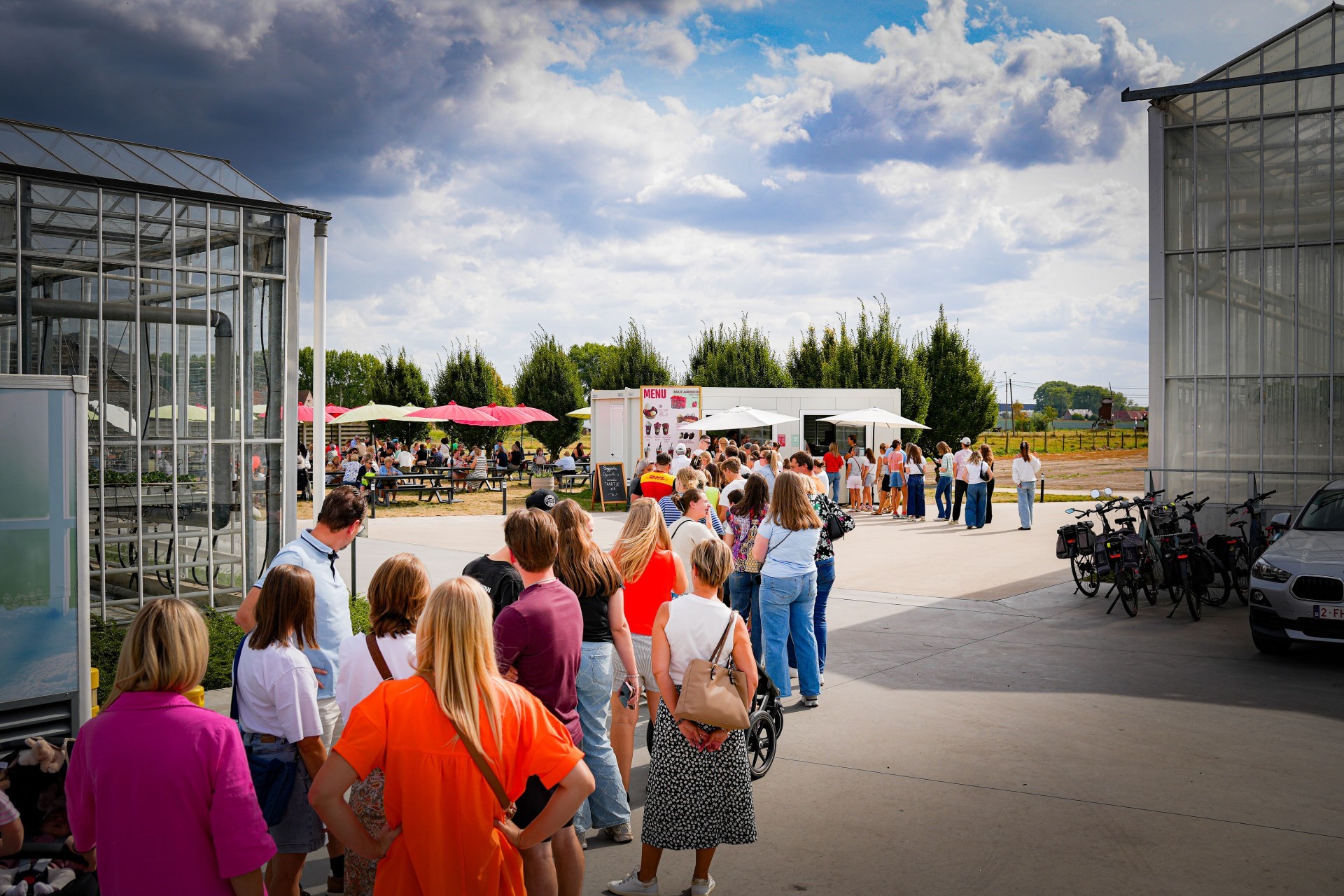 Bezoekers wachten in de rij tussen twee serres van de boerderij in Meulebeke onder een dramatische zomerhemel.